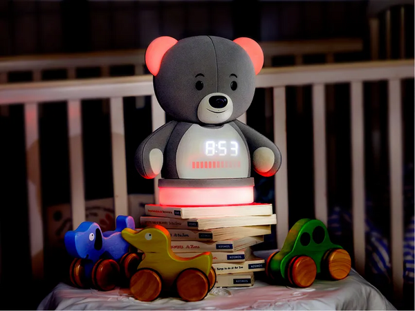 DreamCub gray bear baby monitor glowing pink with a clock on its chest display, beside a crib with wooden toy cars